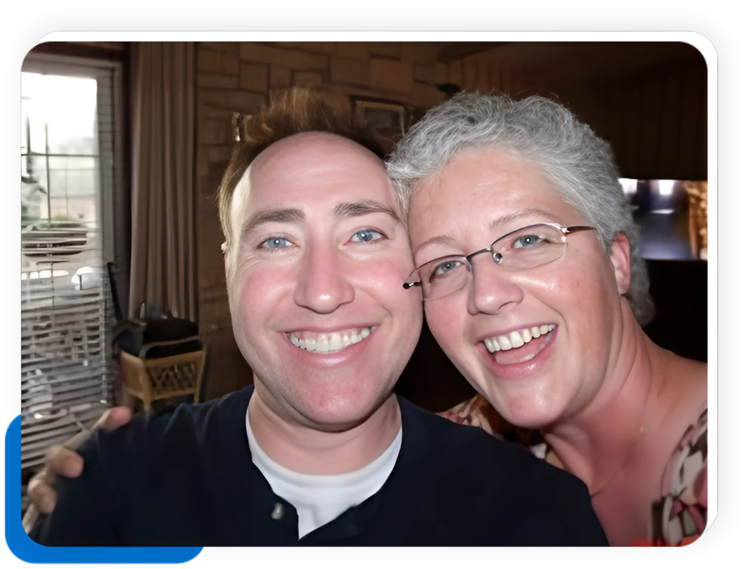 A man and woman, both smiling widely, take a close-up selfie indoors. The man, in a dark shirt and with short hair, stands next to the woman, who has short gray hair and glasses about her face, wearing a lovely floral top.